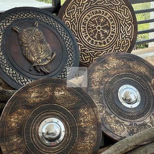 May include: Four intricately carved wooden shields. One features an owl, another a compass design, and two with circular patterns and metal centerpieces. The shields are brown with gold and silver accents, showcasing detailed craftsmanship.