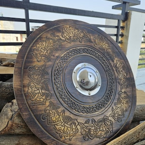 May include: A round, wooden shield with intricate carved designs and a central metal boss. The shield features a dark brown stain, with detailed knotwork patterns around the edges and center. The shield is approximately 30 inches in diameter.