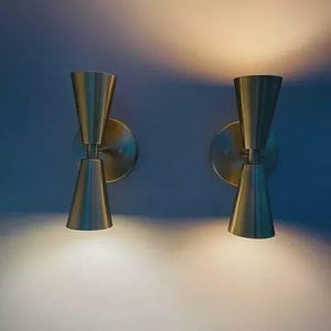 Peut inclure: Deux appliques murales modernes avec une finition en métal brossé. Chaque applique est dotée d'un abat-jour conique et d'une plaque arrière circulaire. Les appliques sont fixées sur un mur bleu, avec une lumière chaude émanant des luminaires.