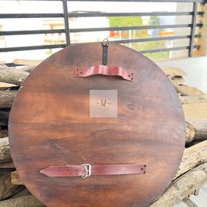 May include: A round, wooden shield with a dark brown finish. It features two red leather straps and a handle on the back for grip and support. The shield is designed for display or costume use.