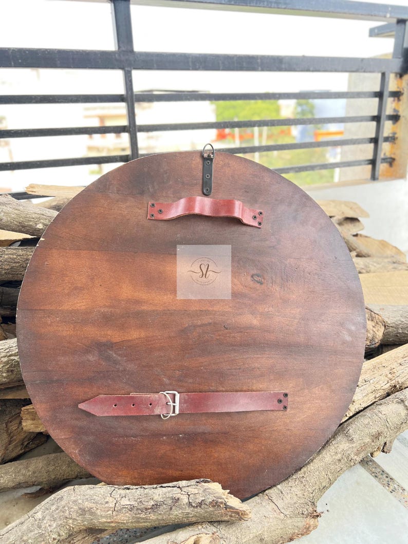 May include: A round, wooden shield with a dark brown finish. It features two red leather straps and a handle on the back for grip and support. The shield is designed for display or costume use.