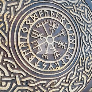 May include: A round, wooden carving with a detailed, etched design. The central focus is a compass-like symbol surrounded by runic text and intricate knotwork patterns. The wood has a dark brown stain with lighter, natural wood tones in the carved areas.