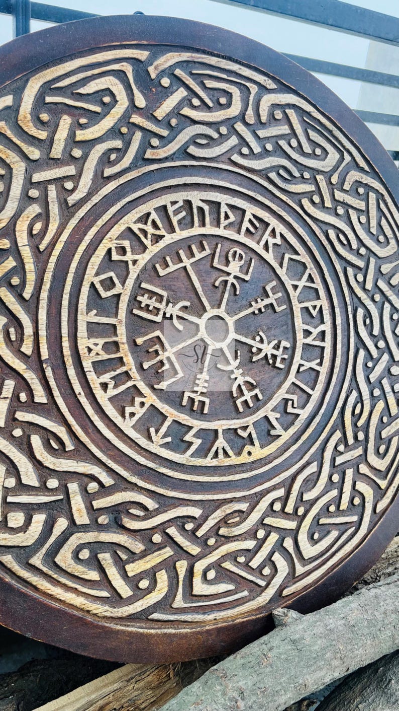 May include: A round, wooden carving with a detailed, etched design. The central focus is a compass-like symbol surrounded by runic text and intricate knotwork patterns. The wood has a dark brown stain with lighter, natural wood tones in the carved areas.