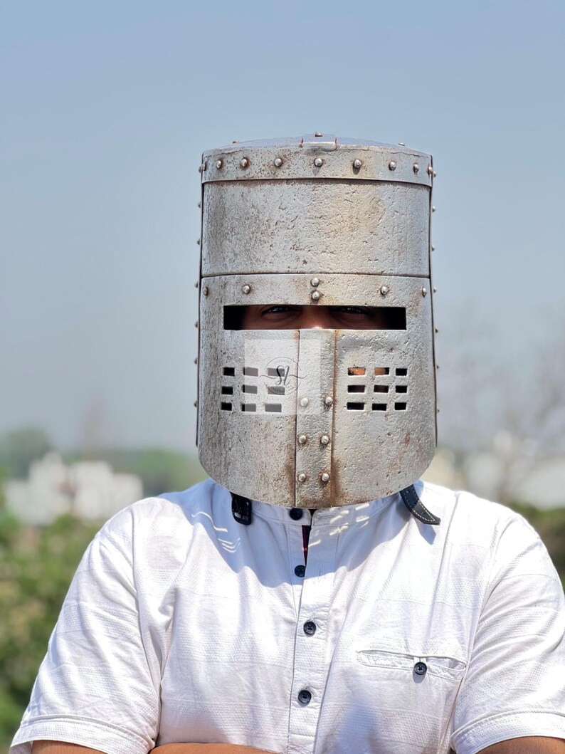 K&ouml;nnte beinhalten: Ein silberfarbener, genieteter Ritterhelm mit rechteckigen Augen- und Bel&uuml;ftungsschlitzen. Der Helm wird &uuml;ber einem wei&szlig;en Hemd mit Kragen getragen. Der Helm hat eine zylindrische Form und ein verwittertes Aussehen.