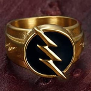 May include: A gold-colored ring featuring a black circular emblem with a lightning bolt design. The ring has a wide band and is set against a dark, textured background.