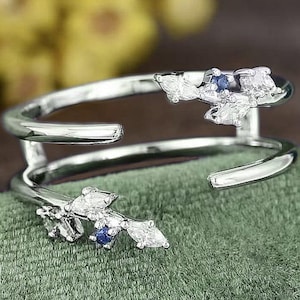 May include: A silver ring with a delicate design featuring a cluster of small white and blue gemstones. The ring is double-banded and has a unique, asymmetrical shape.