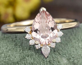 May include: A gold ring featuring a pear-shaped, light pink gemstone as the centerpiece. The ring is adorned with small, round, clear gemstones surrounding the main stone and a single pear-shaped stone below. The ring is set on a green surface.