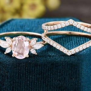 K&ouml;nnte beinhalten: Zwei ros&eacute;goldene Ringe. Ein Ring hat einen ovalen, hellrosa Edelstein, der von marquisef&ouml;rmigen Steinen umgeben ist. Der zweite Ring hat ein Chevron-Design, das mit kleinen, klaren Edelsteinen besetzt ist. Beide Ringe liegen auf einem dunkelblauen Samtstoff.