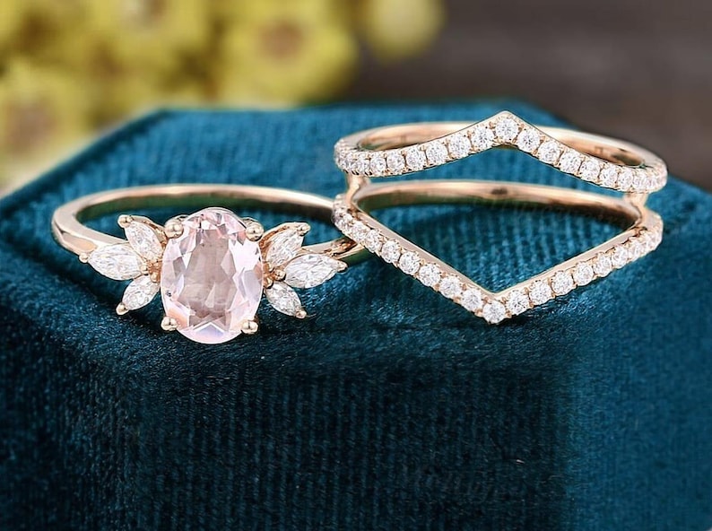 K&ouml;nnte beinhalten: Zwei ros&eacute;goldene Ringe. Ein Ring hat einen ovalen, hellrosa Edelstein, der von marquisef&ouml;rmigen Steinen umgeben ist. Der zweite Ring hat ein Chevron-Design, das mit kleinen, klaren Edelsteinen besetzt ist. Beide Ringe liegen auf einem dunkelblauen Samtstoff.