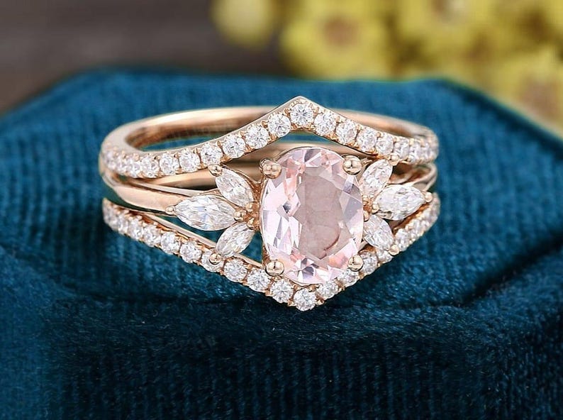 K&ouml;nnte beinhalten: Ein Verlobungsring-Set aus Ros&eacute;gold mit einem oval geschliffenen Morganit-Mittelstein und einem diamantbesetzten Band. Der Ring wird von zwei zus&auml;tzlichen B&auml;ndern begleitet, eines mit Chevron-Design und das andere mit einem Diamantband.