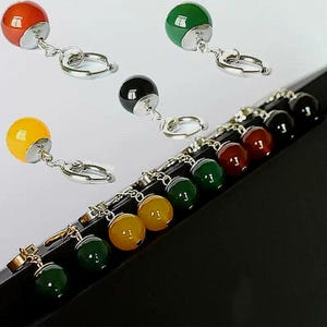 Könnte beinhalten: Eine Sammlung farbenfroher Billardkugel-Schlüsselanhänger. Die Schlüsselanhänger zeigen verschiedenfarbige Kugeln, darunter rot, grün, gelb und schwarz, die an silberfarbenen Metallringen befestigt sind. Die Schlüsselanhänger werden auf einer schwarzen Oberfläche präsentiert.