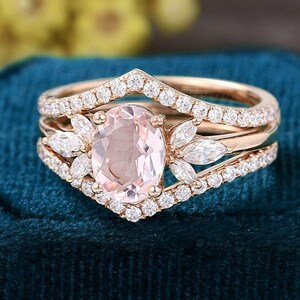 K&ouml;nnte beinhalten: Ein Verlobungsring-Set aus Ros&eacute;gold mit einem oval geschliffenen, hellrosa Edelstein. Der Ring ist von kleineren, klaren Edelsteinen umgeben und hat ein pfeilf&ouml;rmiges Band. Das Set enth&auml;lt zwei zus&auml;tzliche B&auml;nder mit klaren Edelsteinen.