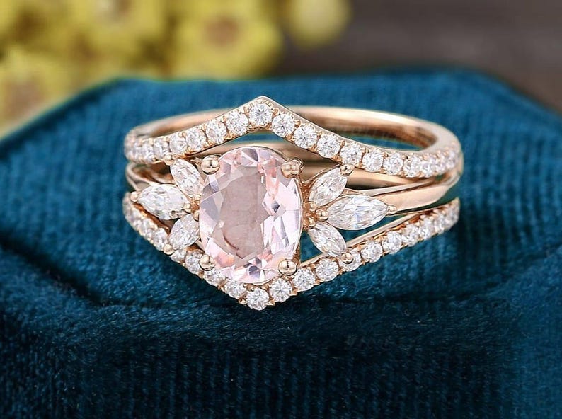 K&ouml;nnte beinhalten: Ein Verlobungsring-Set aus Ros&eacute;gold mit einem oval geschliffenen, hellrosa Edelstein. Der Ring ist von kleineren, klaren Edelsteinen umgeben und hat ein pfeilf&ouml;rmiges Band. Das Set enth&auml;lt zwei zus&auml;tzliche B&auml;nder mit klaren Edelsteinen.