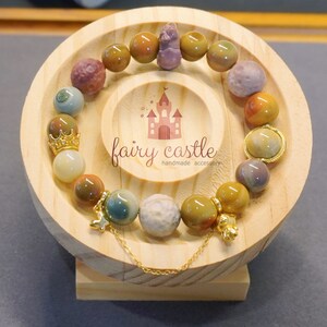 May include: A multi-coloured gemstone bracelet with gold accents and charms. The bracelet is displayed on a wooden stand with the text "fairy castle handmade accessory".