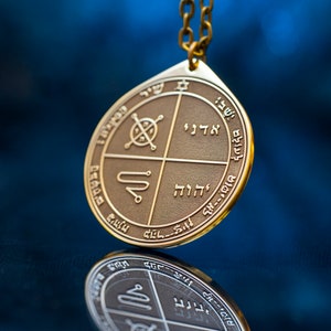 May include: A gold pendant with a circular design featuring Hebrew text and symbols. The pendant is hanging from a gold chain.