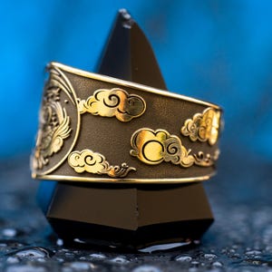 Puede incluir: Un anillo de oro con fondo negro y un dise&ntilde;o de nubes. El anillo est&aacute; sobre un soporte negro en forma de pir&aacute;mide.