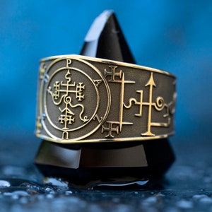 Peut inclure: Une bague en argent sur un fond noir. La bague présente un motif complexe avec un cercle au centre et une croix à l'intérieur du cercle. La bague est entourée d'une série de symboles et de lettres.