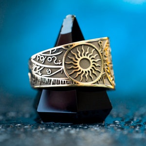 May include: A gold ring with a sun design and intricate engravings. The ring is displayed on a black triangular stand.