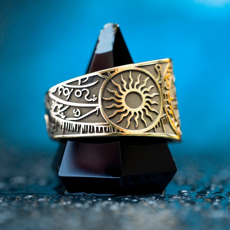 May include: A gold ring with a sun design and intricate engravings. The ring is displayed on a black triangular stand.