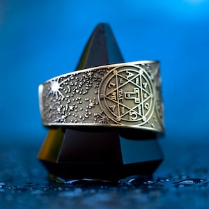 May include: A silver ring with a star-shaped design and a circular inscription in Hebrew. The ring is on a black pyramid-shaped stand against a blue background.