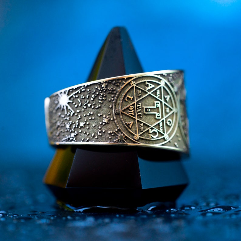 May include: A silver ring with a star-shaped design and a circular inscription in Hebrew. The ring is on a black pyramid-shaped stand against a blue background.