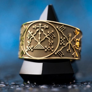 Puede incluir: Un anillo dorado con un diseño intrincado que presenta una cruz, serpientes y estrellas. El anillo está en un soporte de exhibición blanco y negro.