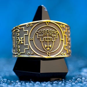 Op de afbeelding: Een goudkleurige ring met ingewikkelde reliëfontwerpen, waaronder symbolen en de letters V, E en N. De ring wordt getoond op een zwarte, geometrische standaard tegen een blauwe achtergrond. Het ontwerp van de ring heeft een centraal embleem en decoratieve randen.
