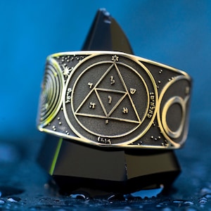 Pode incluir: Um anel de prata com um design de esmalte preto que apresenta um triângulo com letras hebraicas dentro. O anel é cercado por um círculo com uma estrela de David e uma lua crescente.