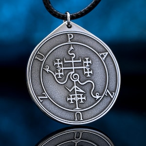 Pode incluir: Um colar com um pingente de prata de design circular que apresenta um símbolo complexo com uma cruz e uma cobra. O pingente é inscrito com as letras "PAZUN" ao redor da borda.