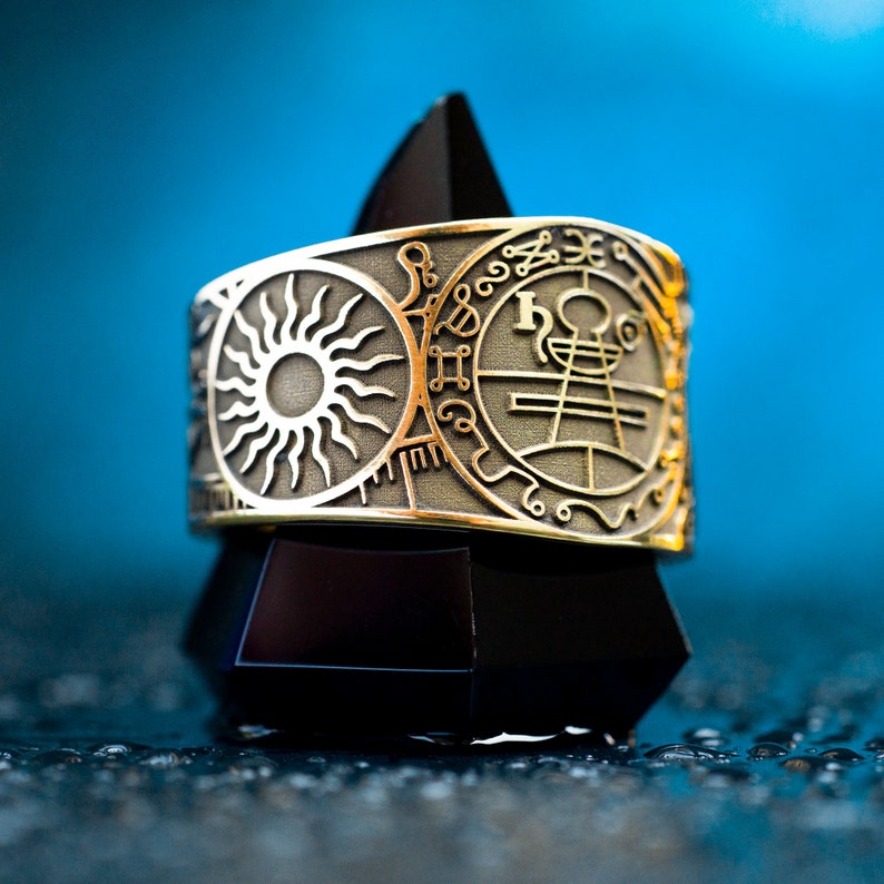 May include: A gold ring with a sun and a circle of symbols on it. The ring is on a black stand against a blue background.