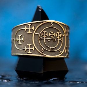 May include: A gold ring with a detailed design featuring a circle with a symbol inside and crosses around the edge. The ring is sitting on a black pyramid-shaped object.