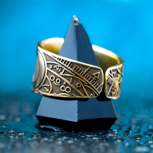 May include: A gold ring with intricate engraved designs, featuring geometric patterns and symbols. The ring is displayed on a black triangular stand against a blue background.