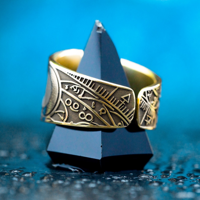 May include: A gold ring with intricate engraved designs, featuring geometric patterns and symbols. The ring is displayed on a black triangular stand against a blue background.