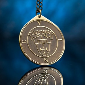 Op de afbeelding: Een gouden hanger met een zegelontwerp, met een kruis en een kroon, met de letters 'E', 'V', 'I' en 'N' rond de rand.