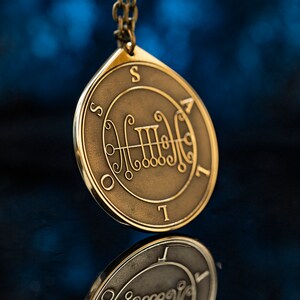 May include: A gold-toned pendant necklace with a circular charm featuring a sigil and the letters "S.O.T.I." around the edge.