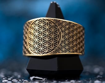 Puede incluir: Un anillo de color dorado con un diseño geométrico detallado, que se asemeja a un patrón de flor de la vida. El anillo se muestra en un soporte negro.