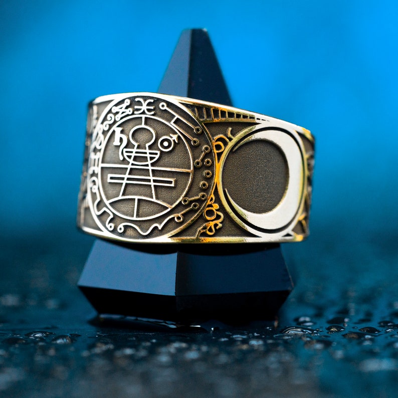 May include: A gold ring with a silver crescent moon and a circular design featuring a symbol and text. The ring is displayed on a black pyramid-shaped stand.