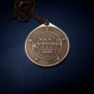 Puede incluir: Un colgante de plata con un acabado de metal marrón oscuro, que presenta un diseño circular con un símbolo y las letras "VRAPED" alrededor del borde.