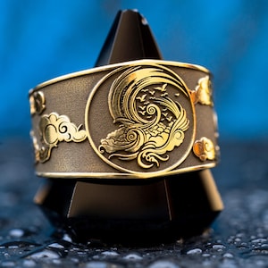 Puede incluir: Un anillo de metal dorado con un dise&ntilde;o detallado de una ola y p&aacute;jaros. El anillo est&aacute; en un soporte negro.