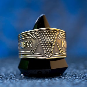 May include: A wide metallic ring with a dark, oxidised finish featuring a triangular engraving of the word ABRACADABRA in a descending pyramid pattern. The band includes additional etched geometric lines and the text SUBTER on the side.