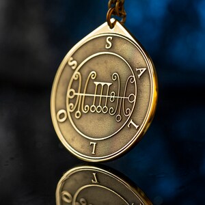 May include: A gold-colored metal pendant with a sigil inscribed on it. The sigil is surrounded by a circle with the letters "S A T I L" inscribed around it. The pendant is hanging from a chain.