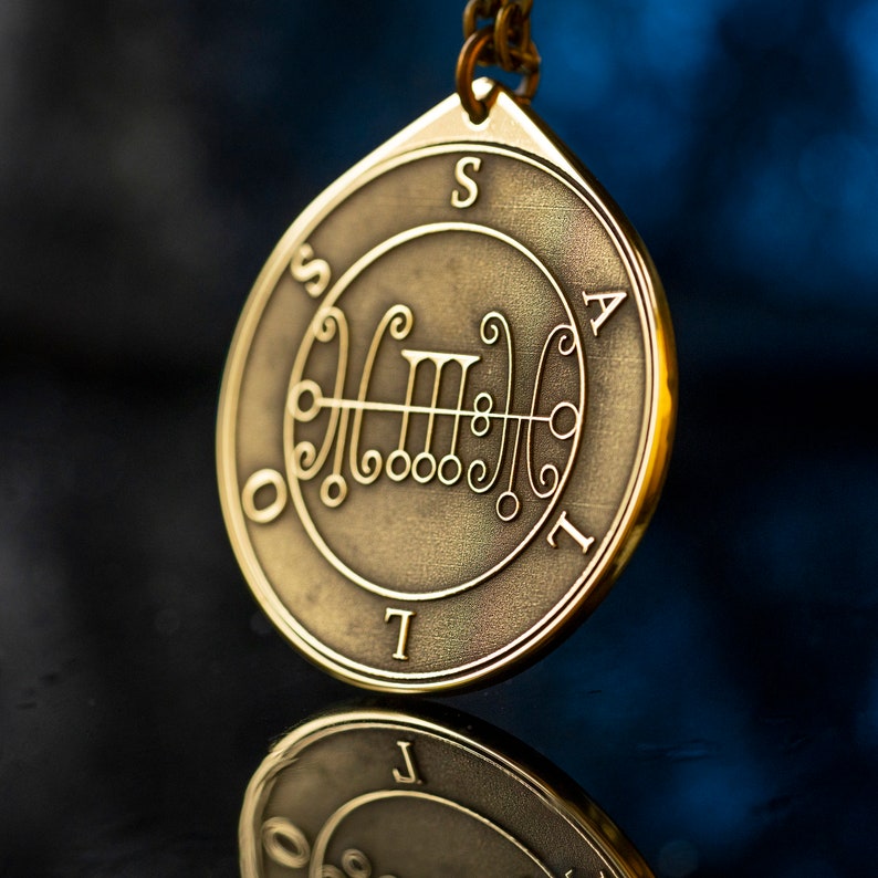 May include: A gold-colored metal pendant with a sigil inscribed on it. The sigil is surrounded by a circle with the letters "S A T I L" inscribed around it. The pendant is hanging from a chain.