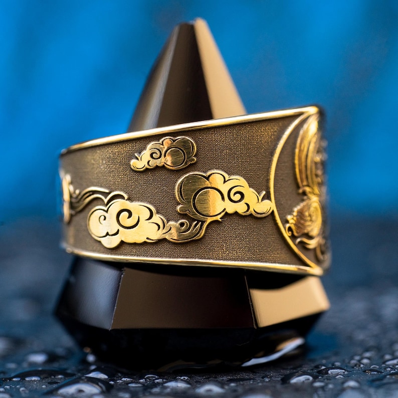 Puede incluir: Un anillo de oro con una banda texturizada de color marr&oacute;n oscuro y un dise&ntilde;o de nubes estilizadas en oro. El anillo se muestra en un soporte c&oacute;nico negro.