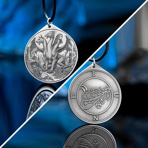 Könnte beinhalten: Ein silberfarbener runder Anhänger an einem schwarzen Band. Eine Seite zeigt einen eingravierten dreiköpfigen Drachen mit Flügeln. Die Rückseite zeigt ein kreisförmiges Siegel mit den Buchstaben B, U, N und E am äußeren Rand der Metallscheibe.