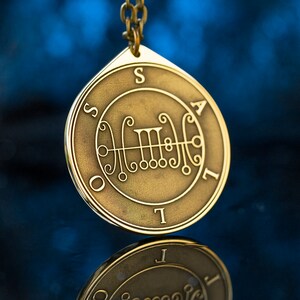 May include: A gold-colored pendant with a sigil and the letters "SAIL" around the edge. The pendant is hanging from a chain.