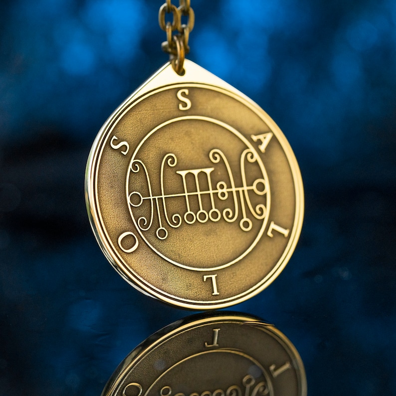 May include: A gold-colored pendant with a sigil and the letters "SAIL" around the edge. The pendant is hanging from a chain.