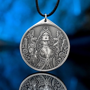May include: A silver pendant necklace with a detailed relief of a woman with long hair, a crown, and snakes. She holds a chalice, and the design includes architectural elements. The pendant hangs from a black cord.
