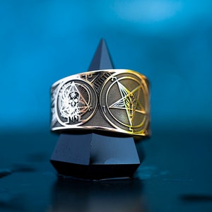 Op de afbeelding: Een zilveren en gouden ring met een pentagram en schedelontwerp. De ring staat op een zwarte piramidevormige standaard.