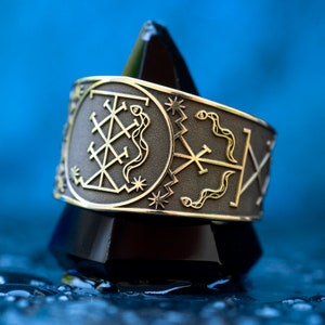 May include: A silver ring with a gold design featuring a geometric pattern and two snakes. The ring is on a black triangular stand against a blue background.