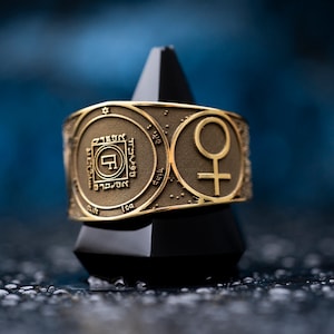 Puede incluir: Un anillo de oro con un diseño circular que presenta texto hebreo y una estrella de David. El anillo también tiene un símbolo de Venus, un círculo con una cruz.